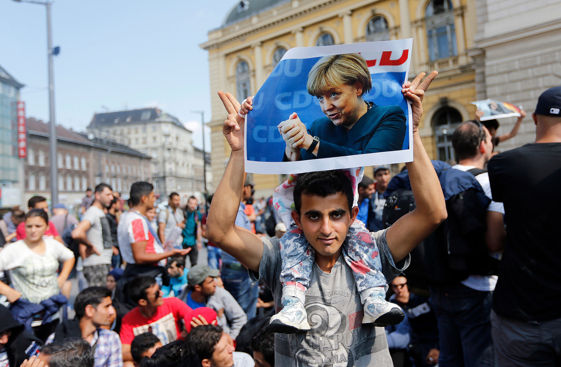 <p>Мигрант демонстрирует портрет Меркель перед тем, как направиться пешком в Австрию из Будапешта, 4 сентября 2015 года</p>

<p>Ангела Меркель была одним из самых заметных сторонников мягкой политики в отношении мигрантов во время кризиса 2015-2017 годов из-за войны в Сирии.&nbsp;&laquo;Мы справимся&raquo;, &ndash;&nbsp;<a href="https://www.politico.eu/article/the-phrase-that-haunts-angela-merkel/" target="_blank">говорила</a>&nbsp;политик про поток мигрантов в Европу. Впоследствии Меркель остро критиковали как за сказанное, так и за послабления в приеме мигрантов, которые, по мнению ее оппонентов, привели к росту преступности со стороны приезжих &mdash; большой резонанс получило, например, <a href="https://tvrain.ru/news/v_germanii-470681/" target="_blank">убийство</a> в Хемнице&nbsp;в 2018 году и <a href="https://www.washingtonpost.com/news/worldviews/wp/2016/07/10/leaked-document-says-2000-men-allegedly-assaulted-1200-german-women-on-new-years-eve/" target="_blank">массовые изнасилования</a> в канун Нового года в декабре 2015 года.</p>